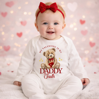 Baby wearing a Valentine-themed onesie with a teddy bear and text, sitting on a soft surface with heart decorations in the background.